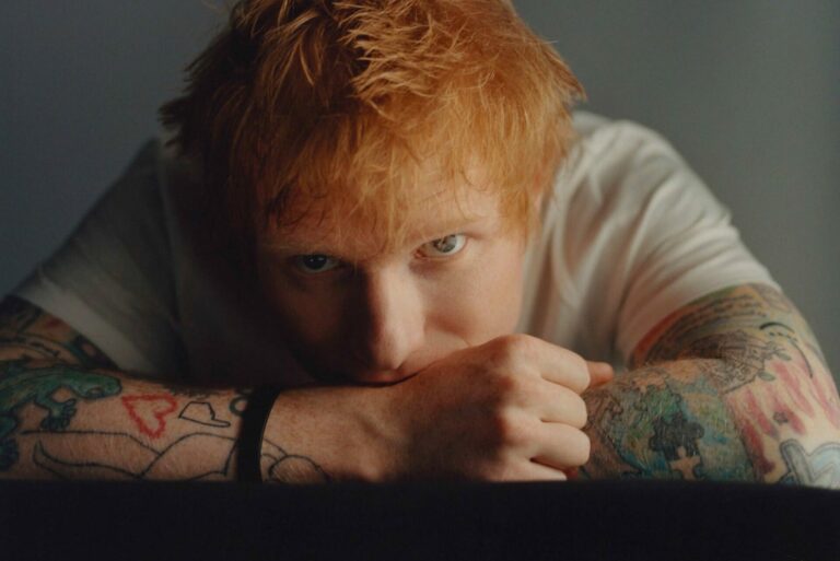 Ed Sheeran poses for a press photo with his head resting on his arms, wearing a white t-shirt