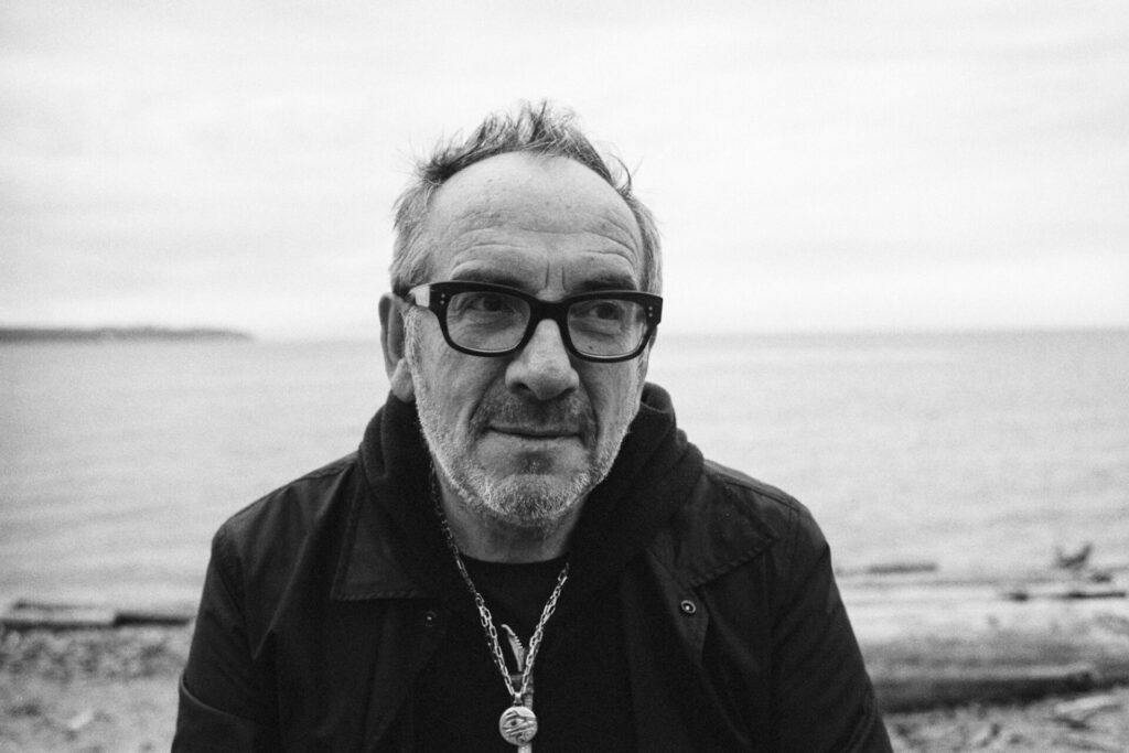 Black and white press photo of Elvis Costello as he poses on a beach