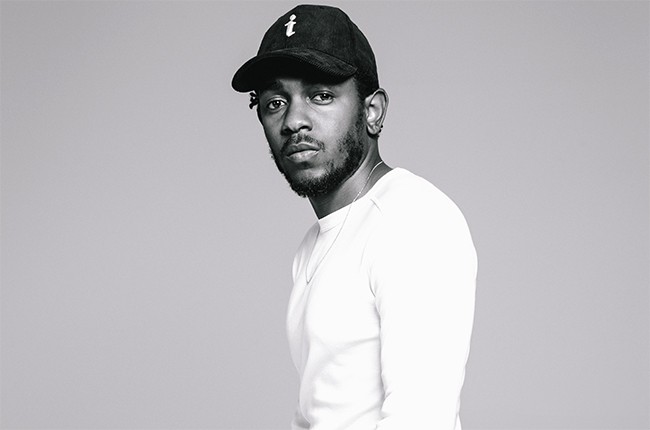 Black and white photo of Kendrick Lamar stood against a plain grey background wearing a white t shirt and black hat