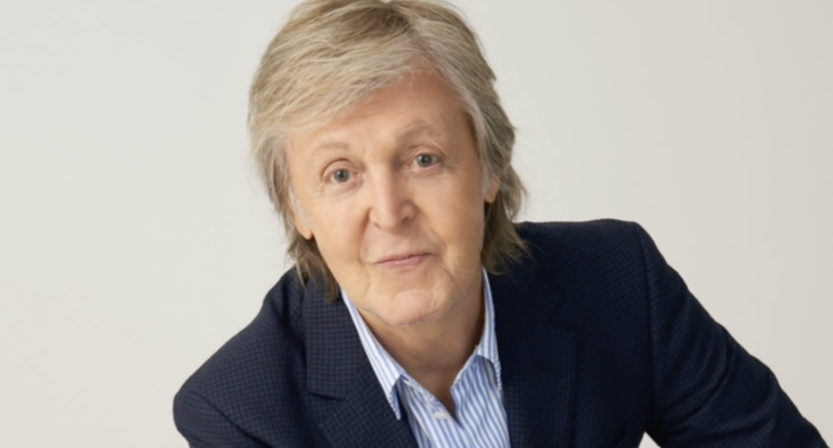 Paul McCartney smiles wearing a suit in front of a white background