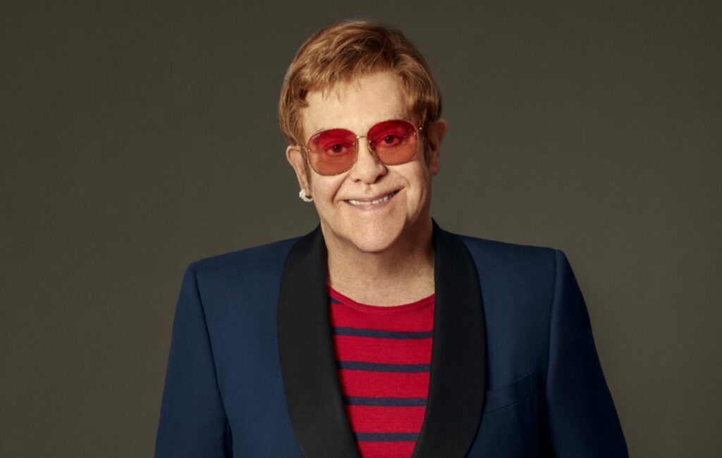 Elton John wears tinted glasses and a blue blazer in a press shot