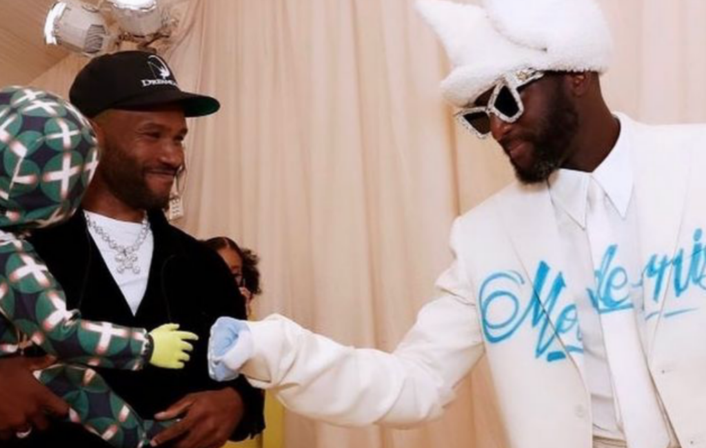 Virgil Abloh wears a white suit and hat and fist bumps Frank Ocean's prop doll on a red carpet