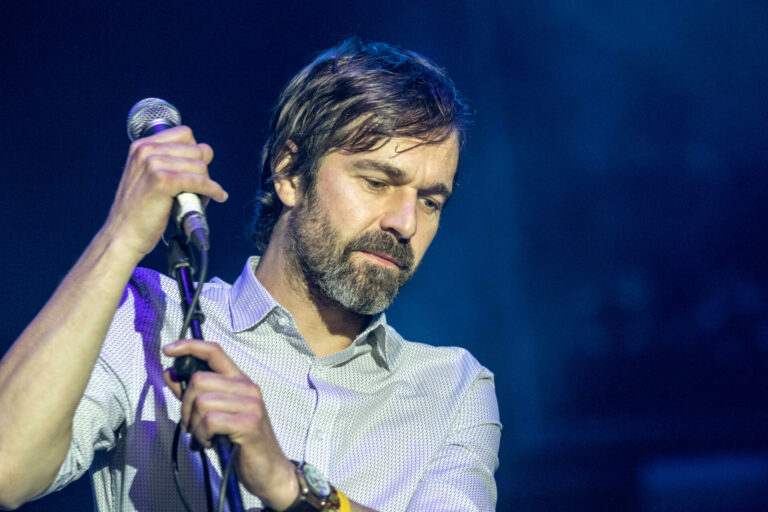 Mark Morriss holds a microphone performing live against a blue background
