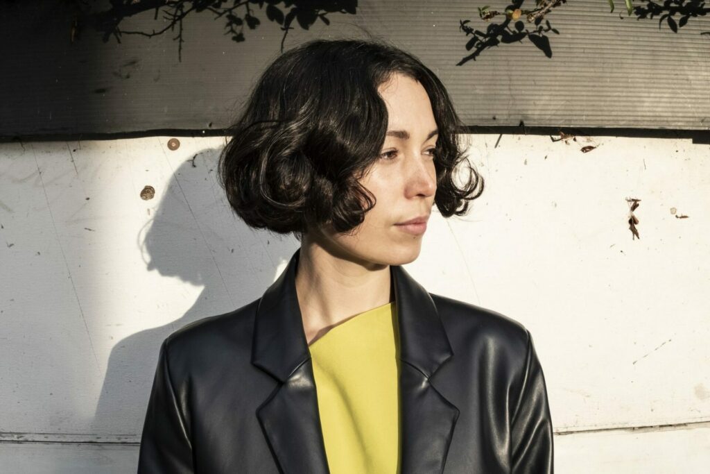Press photo of Kelly Lee Owens turning to one side, wearing a yellow top and a black leather jacket.