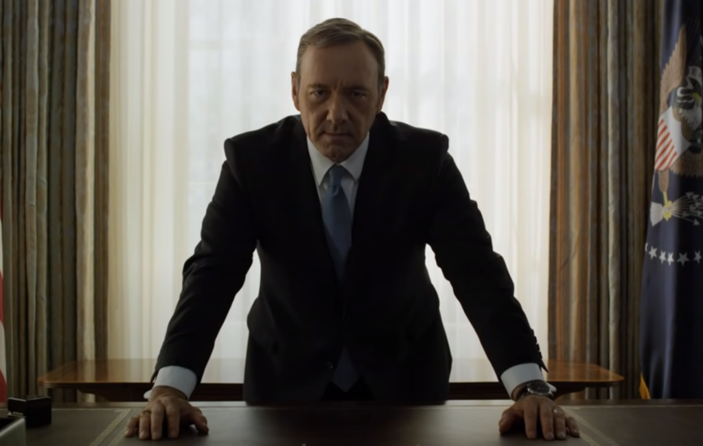 Kevin Spacey wears a suit and leans over a desk playing Frank Underwood in House of Cards