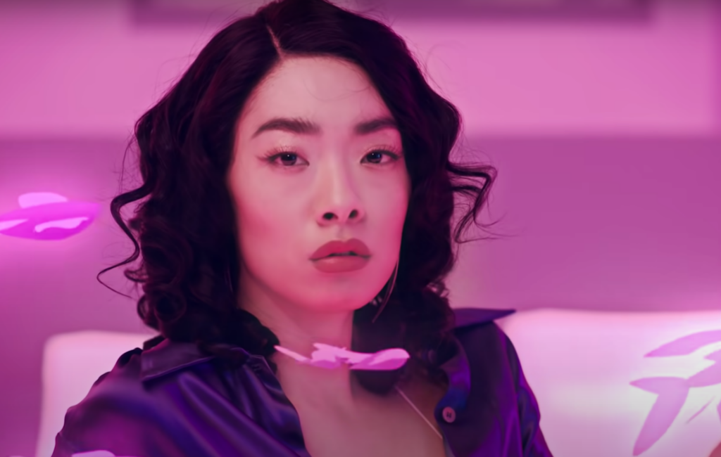 Rina Sawayama wears a pink bow tie and silky blue top, with short, black curled hair