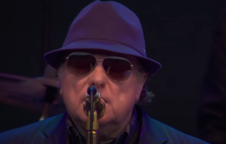 Van Morrison wears a hat and sunglasses performing in front of a microphone in 2017