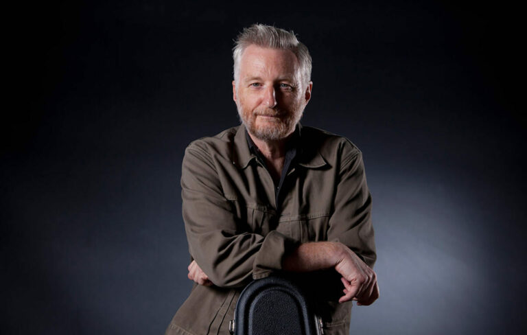 Billy Bragg leaning on a stool and smiling to camera