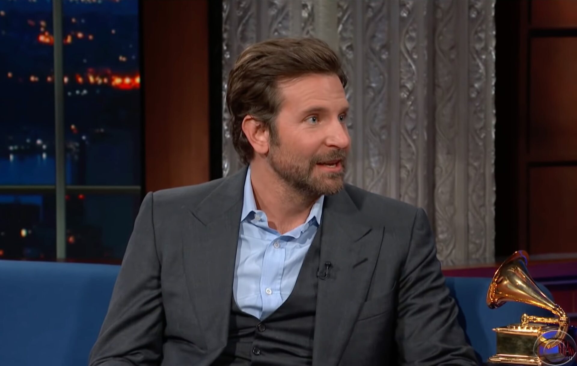 Bradley Cooper sitting on a chat show sofa with a grey suit and blue shirt