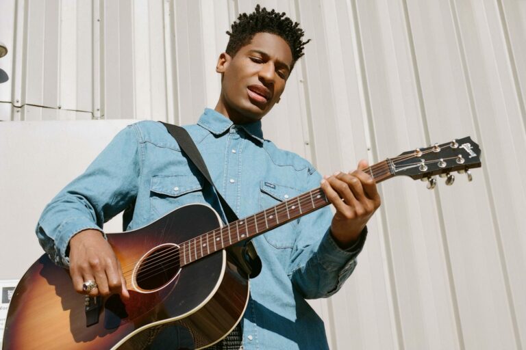 Jon Batiste posing for a press photo holding a guitar and wearing a blue shirt.