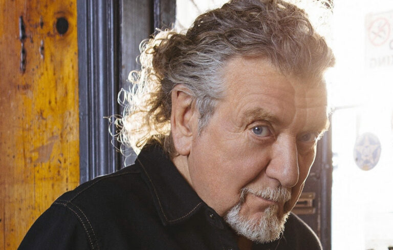 Robert Plant with long hair and a bear, looking sideways at the camera while sunlight comes through a window