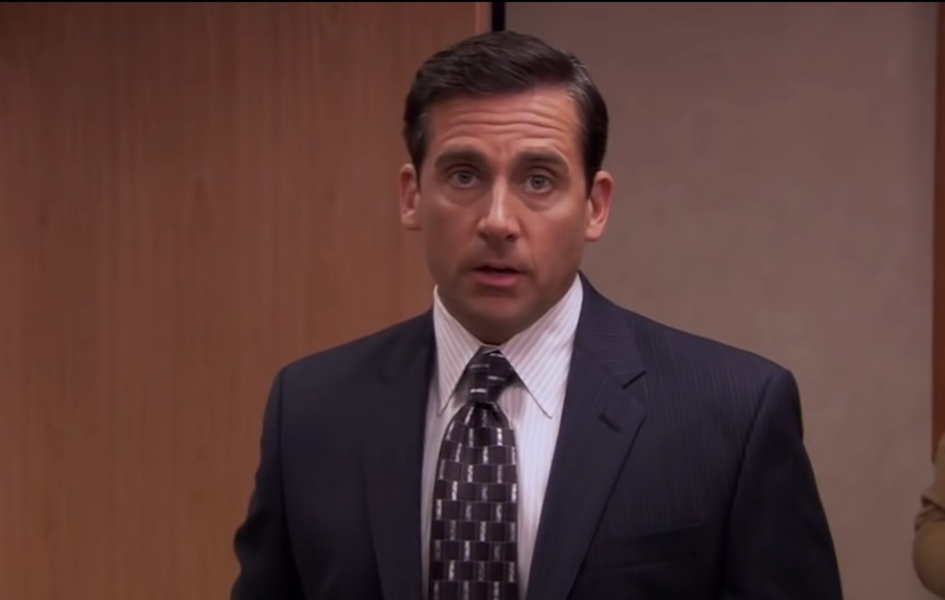 Steve Carell as Michael Scott, wearing a suit against a plain wall