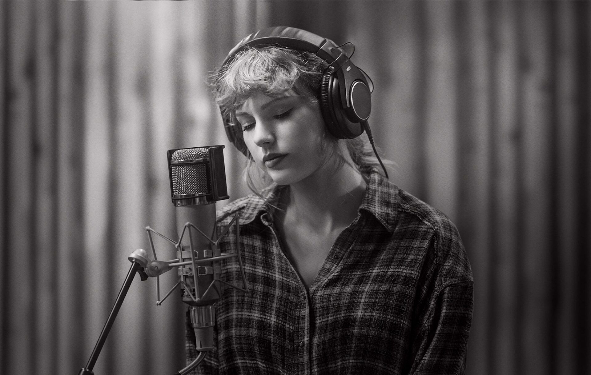 Taylor Swift in black and white looking at the ground wearing headphones in front of a microphone in a recording studio