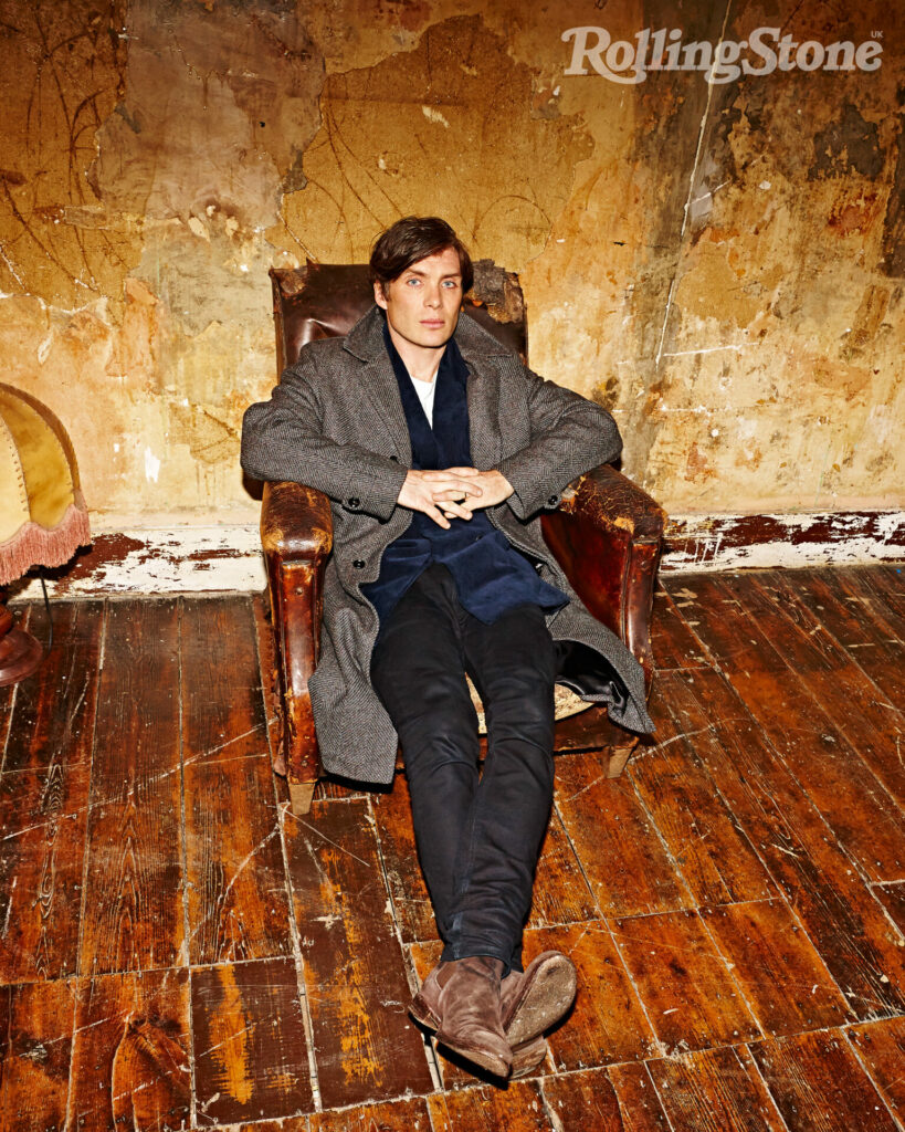 Cillian Murphy sits in a brown leather chair on a hardwood floor against a faded wall
