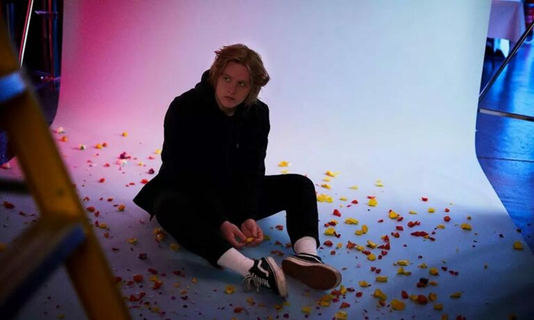Lewis Capaldi poses for a press photo sitting down, surrounding by flower petals