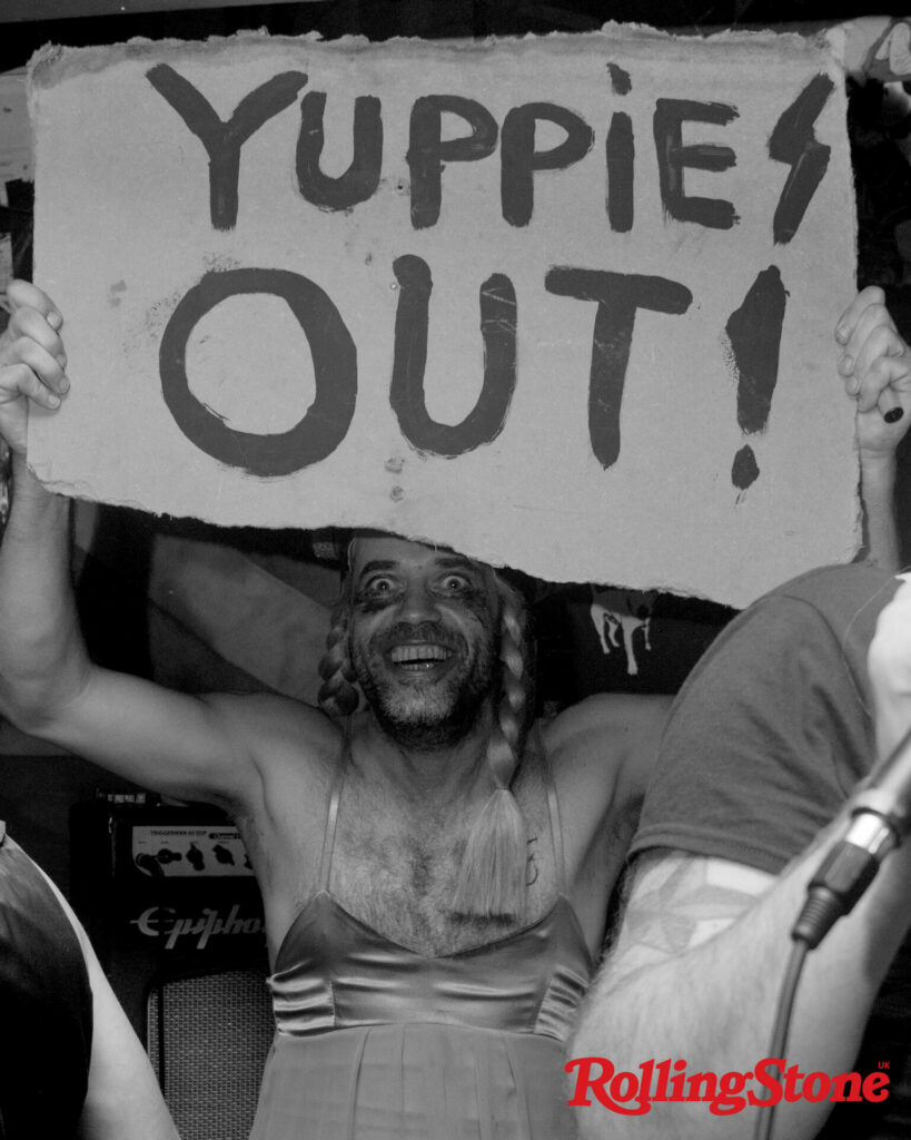 Simon Ticker smiling at the camera wearing a dress and holding a sign reading 'YUPPIES OUT!'
