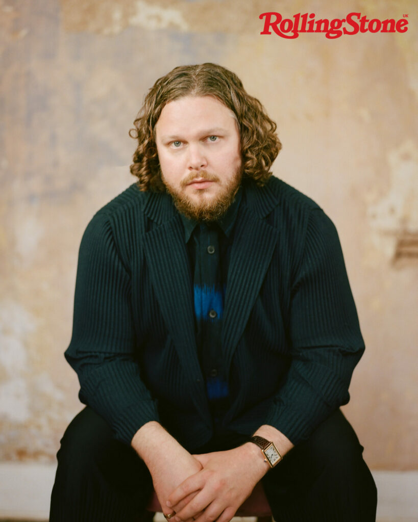 Alt-j's Joe Newman looks at the camera in a navy blue jacket against a faded background