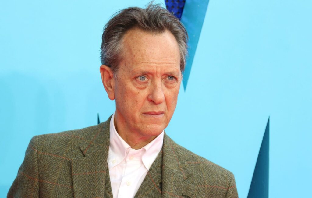 Richard E. Grant poses on a red carpet wearing an olive green suit