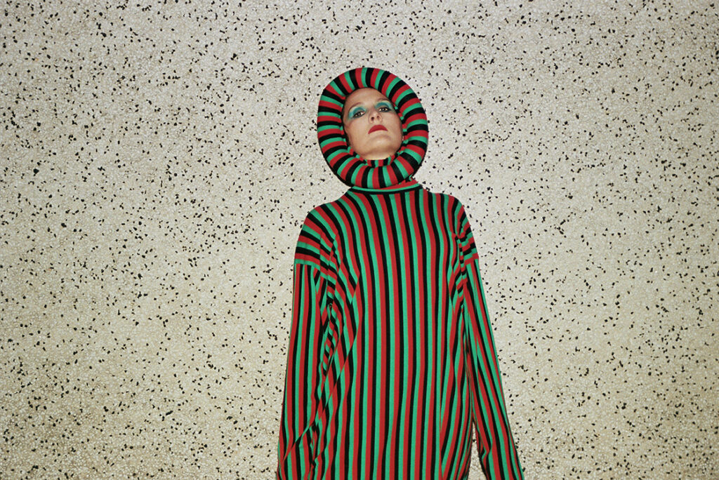 Cate Le Bon wears a striped green, black and red dress with a neckline that wraps around her face