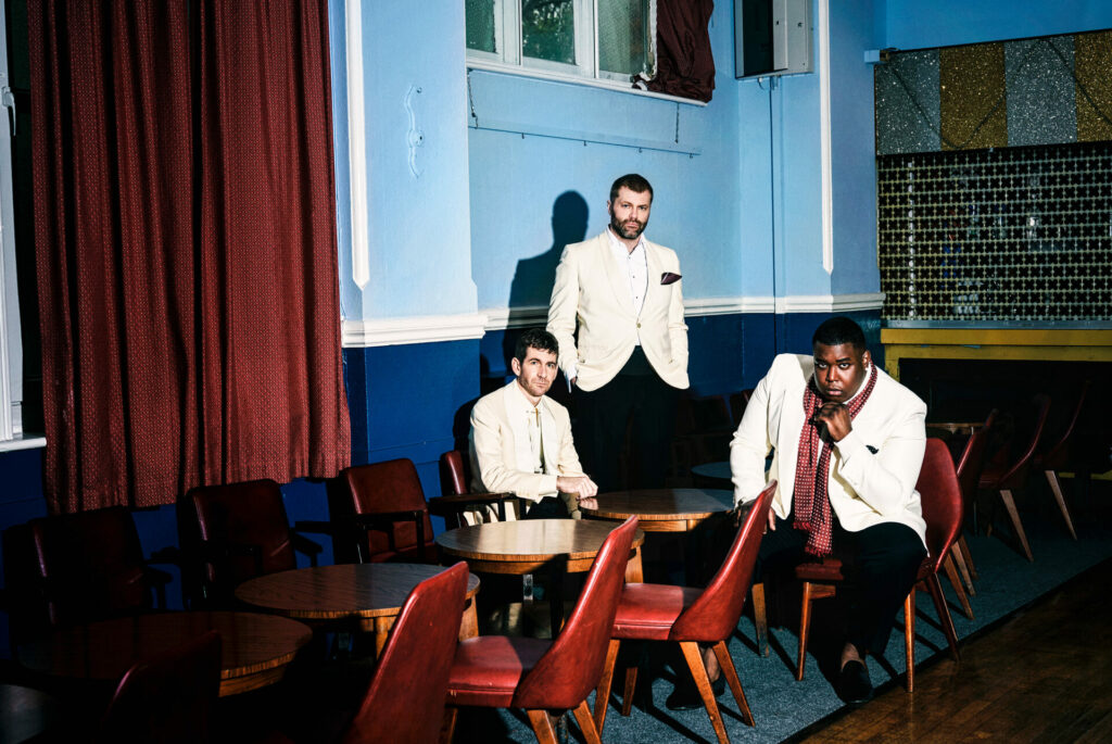 The three members of Gabriels pose on wooden chairs in white suit jackets