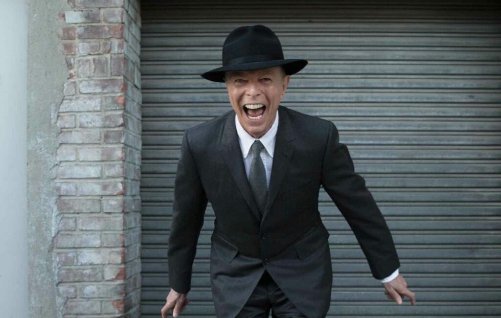 David Bowie wears a suit and a hat standing in front of a garage with a wide grin