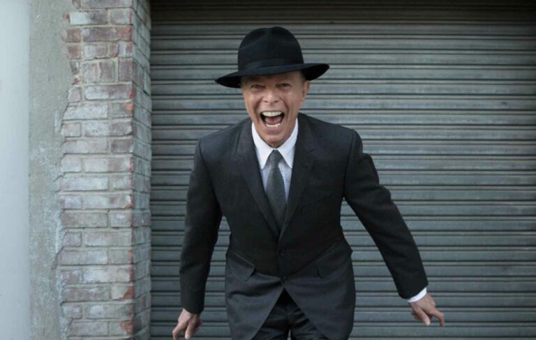 David Bowie wears a suit and a hat standing in front of a garage with a wide grin