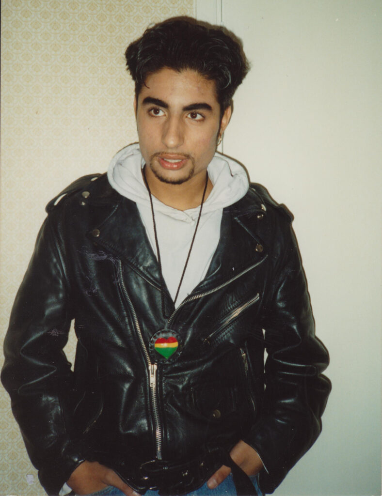 A South Asian man wears a leather jacket, hoodie and heart medallion