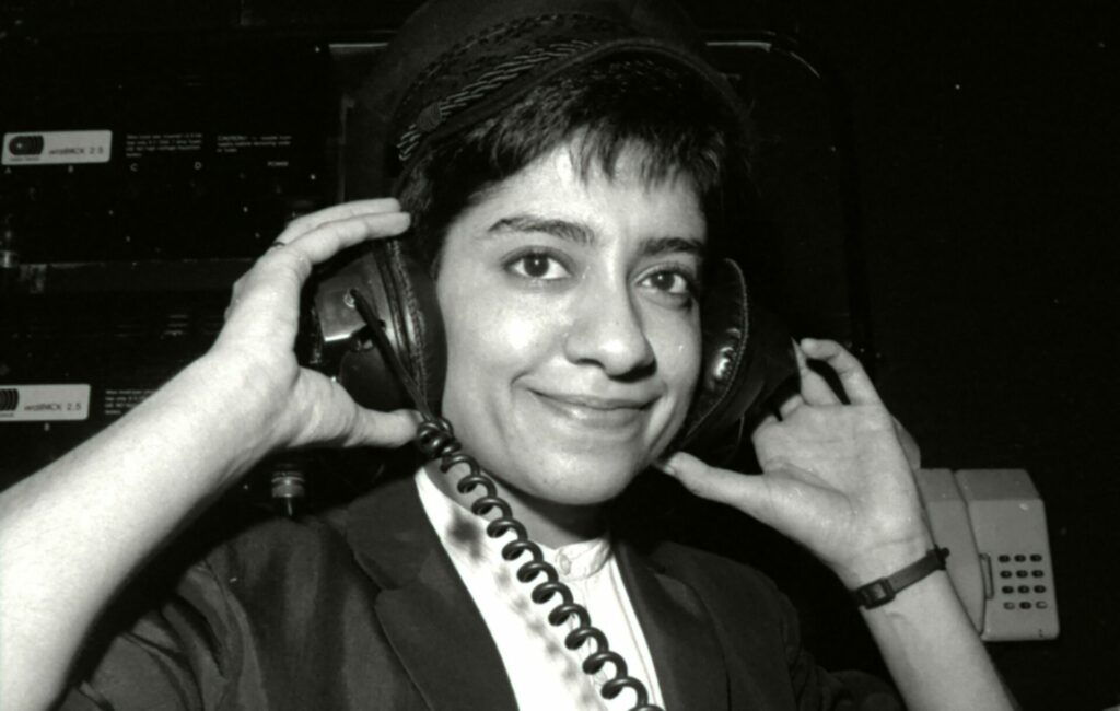 A South Asian DJ wears headphones and smiles at the camera in a close up black and white shot