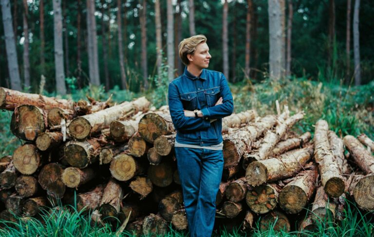 George Ezra poses standing in front of chopped logs in a forest