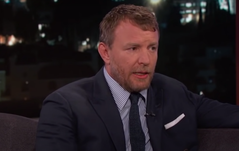 Guy Ritchie wears a navy blue suit and tie on a chat show.