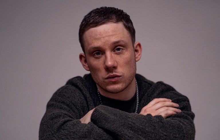 Joe Cole wears a dark green jumper and silver chain, crossing his arms and looking directly at the camera