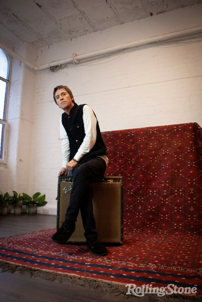 Johnny Marr sits on a guitar amp on a red carpet