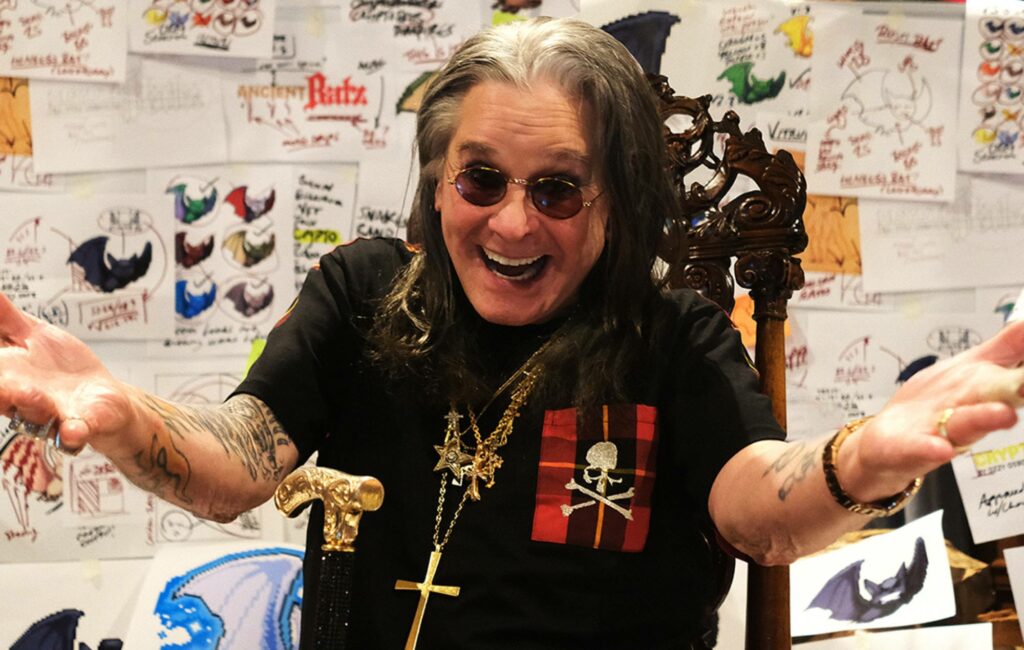 Ozzy Osbourne poses with his hands looking at the camera in black glasses and a black t-shirt