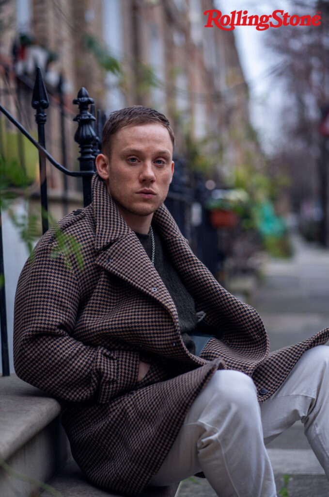 Joe Cole wears a brown and black checkered coat and sits on a doorstep