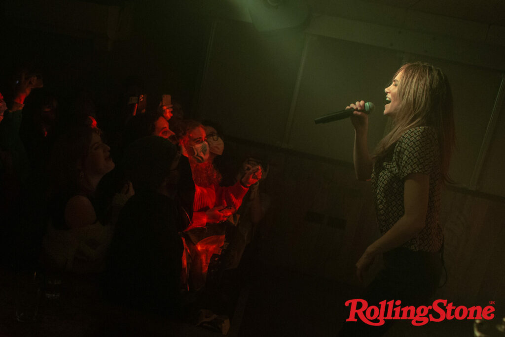 Suki Waterhouse performs live, holding the microphone in a side profile shot