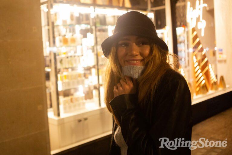 Suki Waterhouse smiles in a grey turtleneck and black jacket and hat outside a shop window