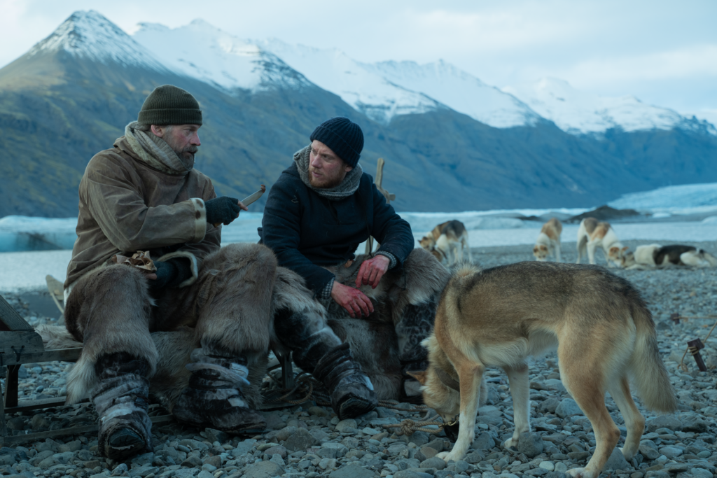 Joe Cole sits in a cold landscape with another man and several dogs, snow capped mountains can be seen in the background