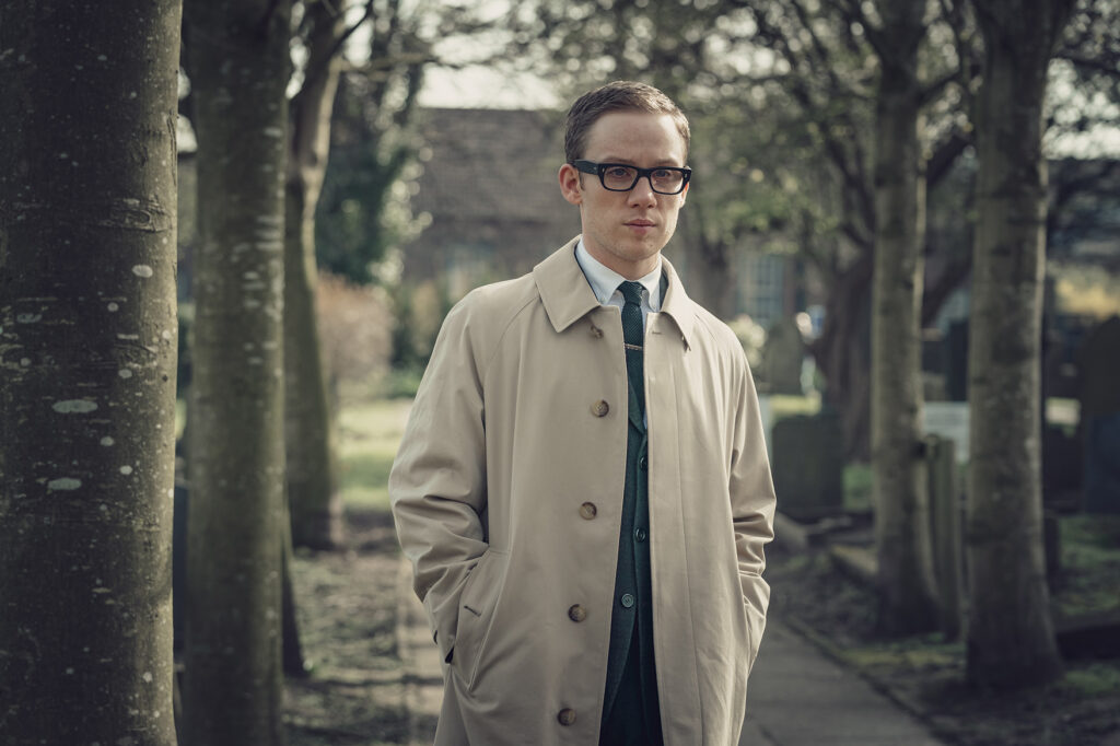 Joe Cole wears a long beige coat and thick black glasses with his hair slicked back