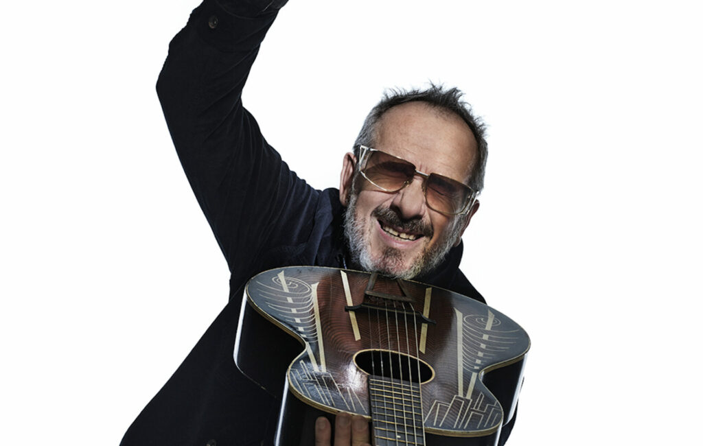 Elvis Costello poses with his head at the end of a guitar pointing towards the camera