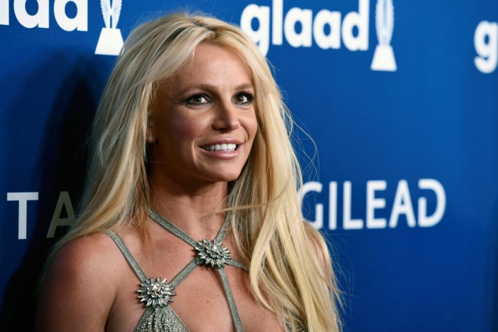 Britney Spears smiles for press against a blue background wearing aa green strappy dress