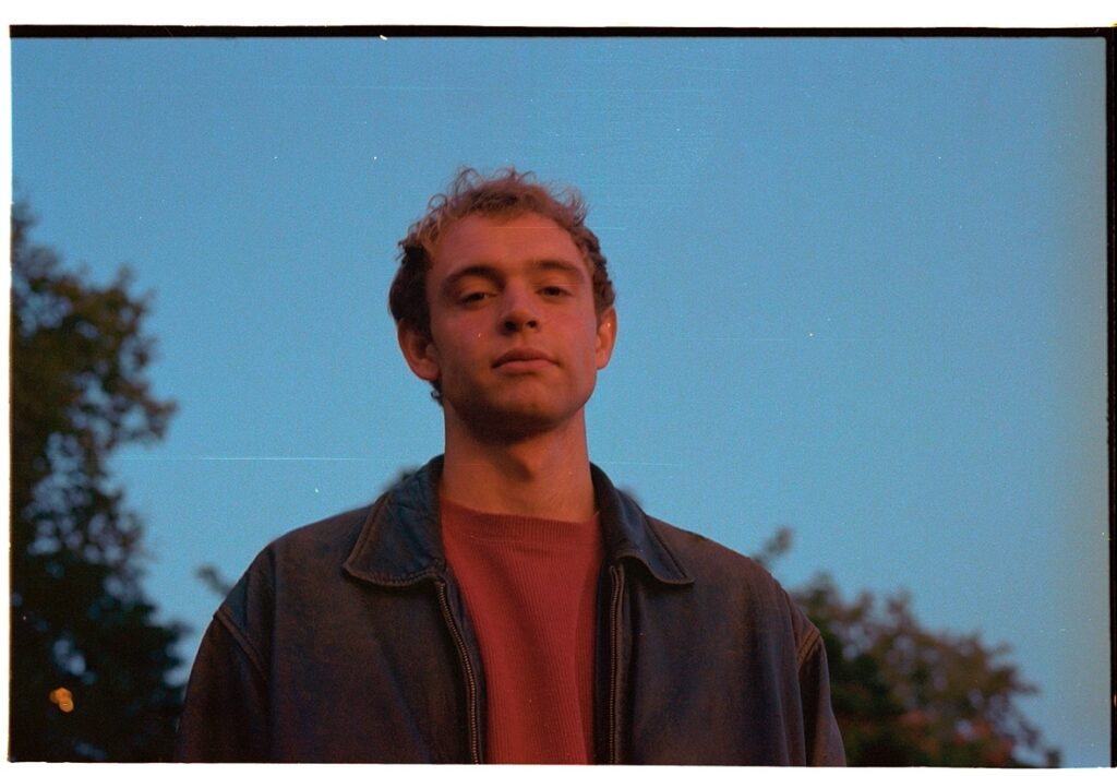 Wunderhorse wears a red t shirt and blue denim jacket in twilight lighting against a blue sky