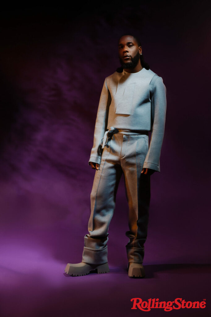 Burna Boy wears a grey outfit and boots in a full body shot against a purple backdrop