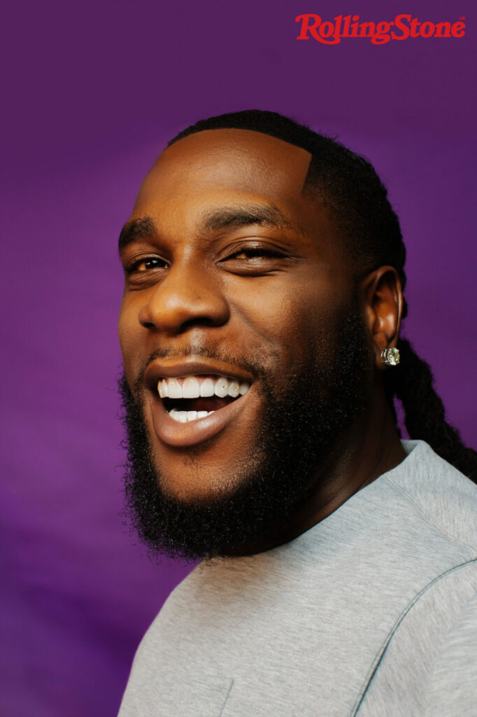 Burna Boy smiles with his teeth in a headshot against a purple backdop