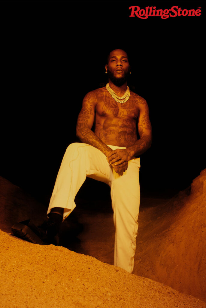 Burna Boy poses shirtless with one leg on a rock structure, wearing white trousers and a diamond chain