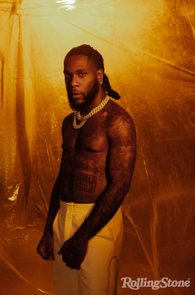 Burna Boy poses shirtless in white trousers in a single angle shot against a plastic backdrop 