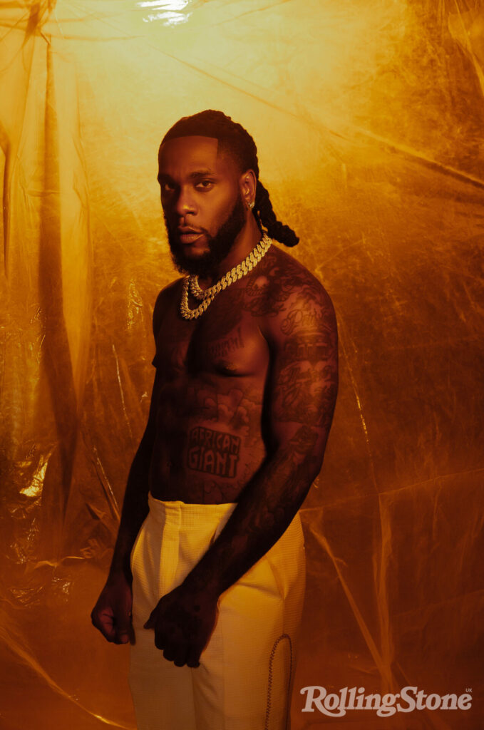 Burna Boy poses shirtless in white trousers in a single angle shot against a plastic backdrop