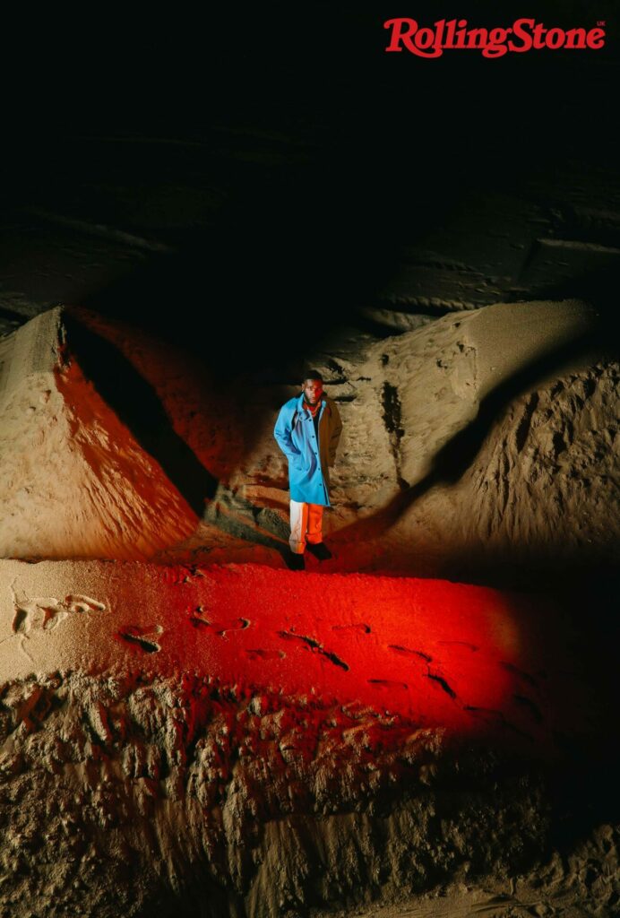 Burna Boy stands at a distance in a cave-like structure wearing a blue jacket