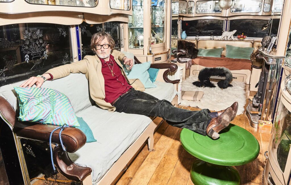Jarvis Cocker poses while reclining on a long sofa with his feet resting on a table
