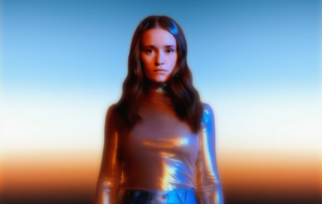 Sigrid wears a silver shimmering shit against an orange cosmic background