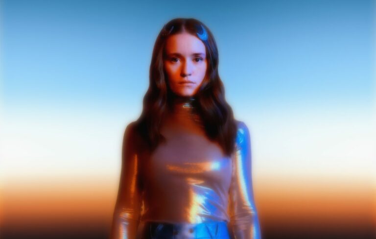 Sigrid wears a silver shimmering shit against an orange cosmic background